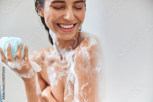 Cheerful young woman with foam on her body holding bath loofah