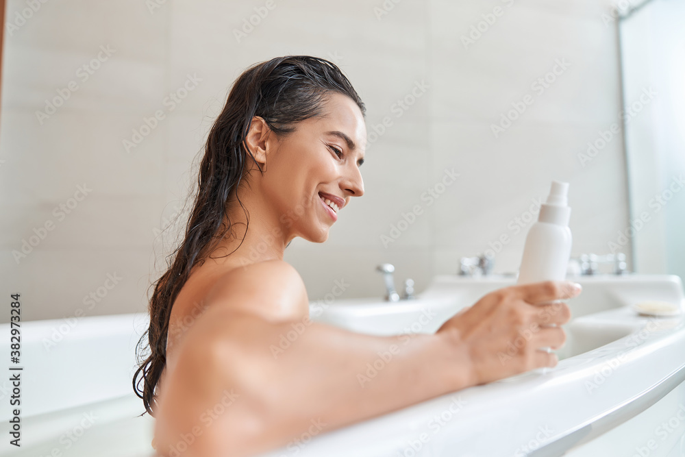 Attractive young woman holding bottle of lotion while relaxing in bathtub Attractive young woman holding bottle of lotion while relaxing in bathtub