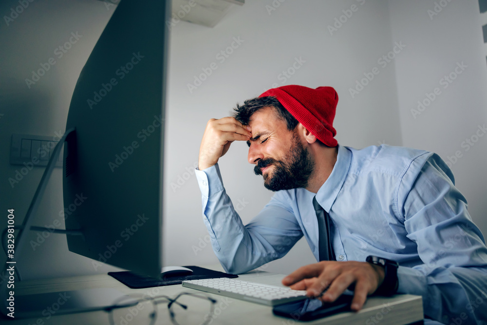 Fototapeta premium Young sad man dressed business casual sitting in his office and holding head. Report is not finished on time.