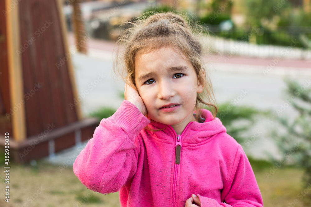A little girl in a pink hoodie with a sad and tearful face is holding her ear. Ear pain, otitis media, swelling of the cheek, gums, toothache, children's surgery, otolaryngology. Children's medicine A little girl in a pink hoodie with a sad and tearful face is holding her ear. Ear pain, otitis media, swelling of the cheek, gums, toothache, children's surgery, otolaryngology. Children's medicine