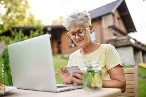 Senior woman with laptop and smartphone working outdoors in garden, home office concept.