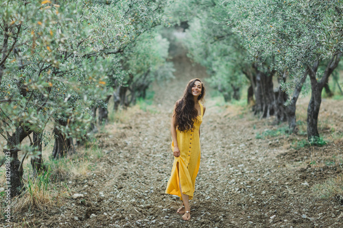 Young happy smiling woman walking in olive tree garden. Yellow linen summer dress. Trendy color and textile style.