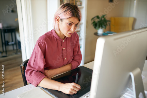 Pretty fashionable student girl with pink hair making illustrations using desktop computer and drawing pad studying online, improving creative skills, having concentrated focused facial expression