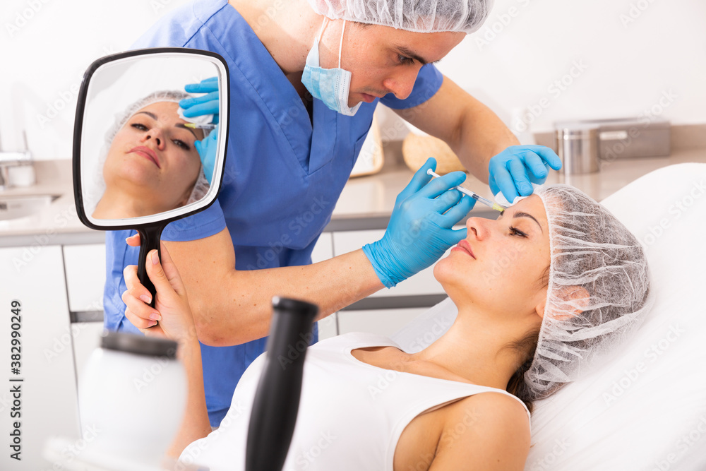 Fototapeta premium Female client looking in mirror during procedure of face mesotherapy in professional clinic of aesthetic cosmetology