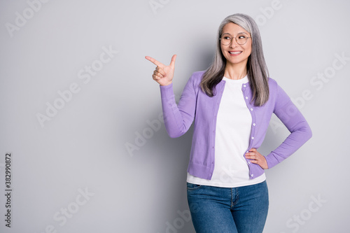 Photo portrait of positive satisfied grandmother smiling looking pointing showing at side with finger wearing jeans violet cardigan spectacles isolated on grey color background