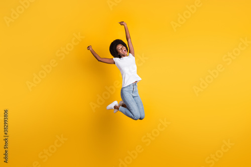 Full length body size photo of funny pretty female millennial with black skin jumping high keeping hands over head isolated on bright yellow color background
