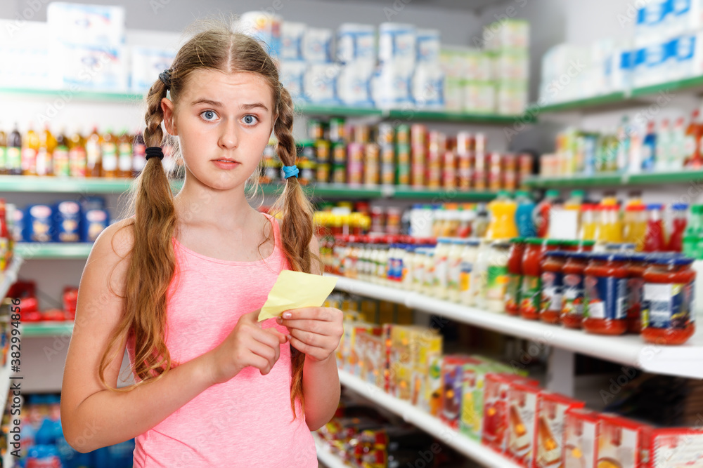 Positive glad cheerful tween girl choosing food products on shopping list in supermarket