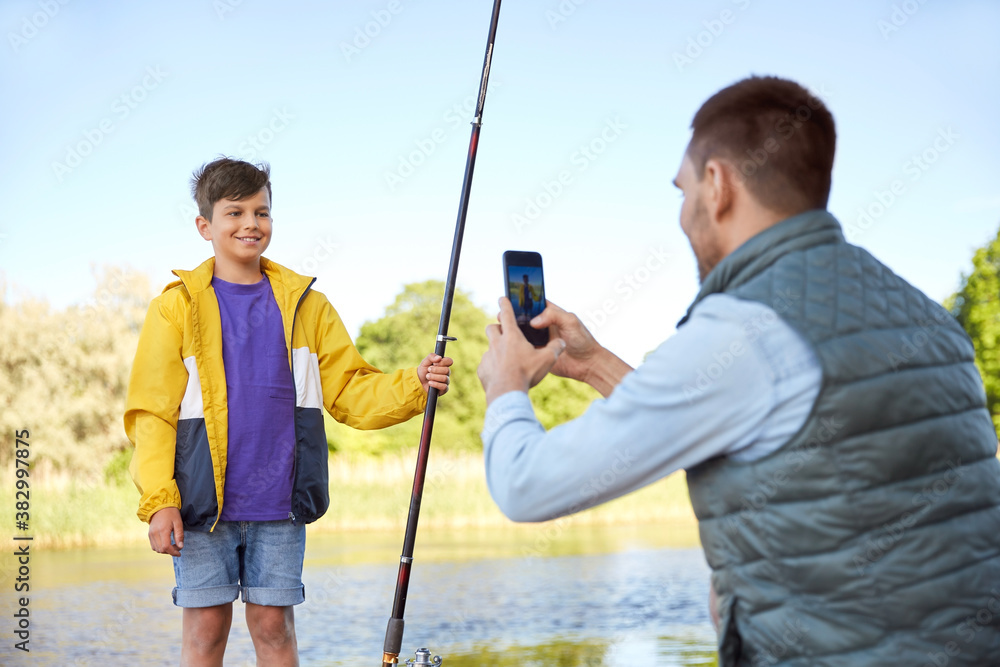 family, generation, summer holidays and people concept - father with smartphone photographing his happy smiling son with fishing rod on river family, generation, summer holidays and people concept - father with smartphone photographing his happy smiling son with fishing rod on river