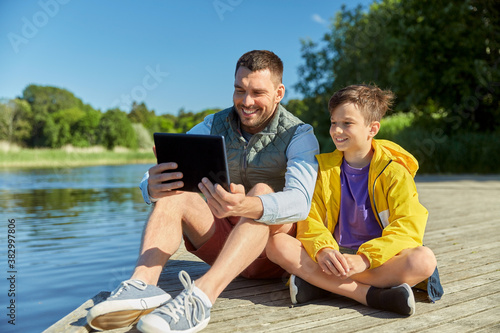 family, generation and technology concept - happy smiling father and son with tablet pc computer sitting on river berth
