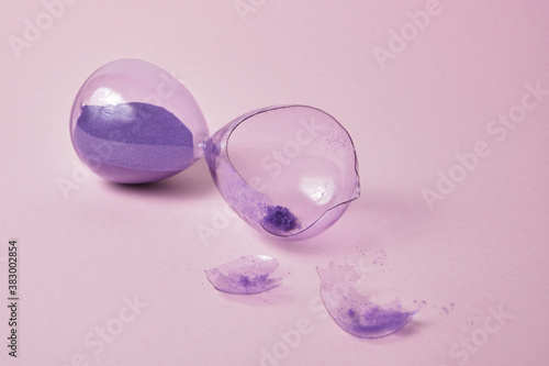 broken glass hourglass with purple sand on a pink background , time concept