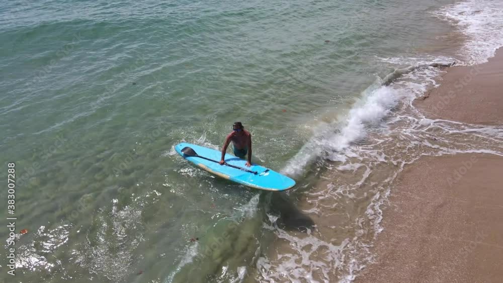 Aerial drone bird's eye view of Caucasian man arrives on beach on a sup paddle board in turquoise tropical clear waters, with beach Aerial drone bird's eye view of Caucasian man arrives on beach on a sup paddle board in turquoise tropical clear waters, with beach