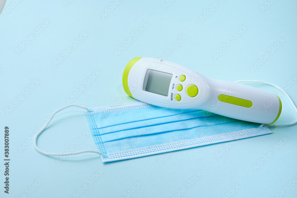 Digital medical infrared non contact thermometer gun for measuring temperature, face mask on blue background Digital medical infrared non contact thermometer gun for measuring temperature, face mask on blue background