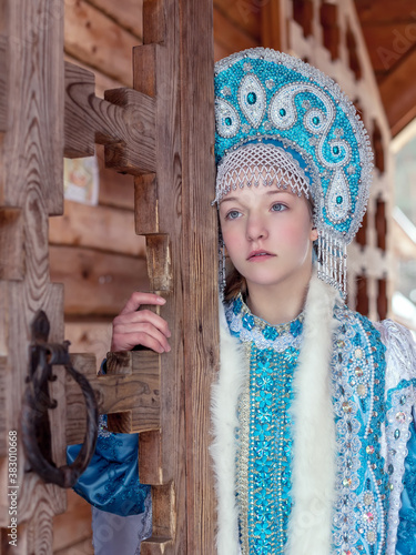 
A girl in a Snow Maiden costume stands against the background of a winter forest and a wooden house. Winter, December. Russian Christmas character Snegurochka. New Year.