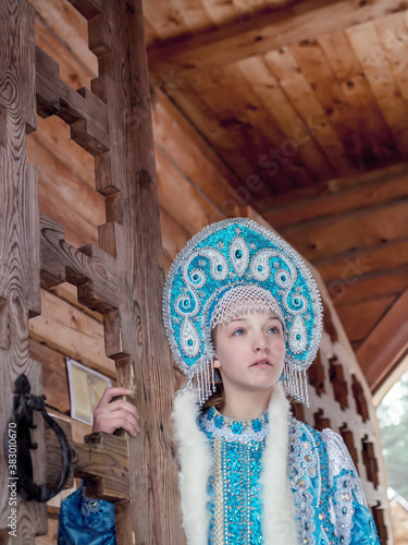
A girl in a Snow Maiden costume stands against the background of a winter forest and a wooden house. Winter, December. Russian Christmas character Snegurochka. New Year.