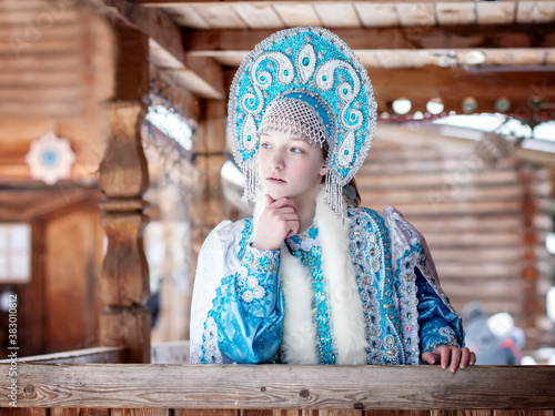 A girl in a Snow Maiden costume stands against the background of a winter forest and a wooden house. Winter, December. Russian Christmas character Snegurochka. New Year.