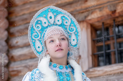 A girl in a Snow Maiden costume stands against the background of a winter forest and a wooden house. Winter, December. Russian Christmas character Snegurochka. New Year.