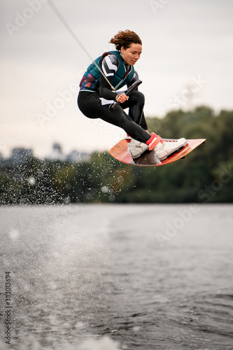 woman in wetsuit holding rope and masterfully jumping over water on wakeboard