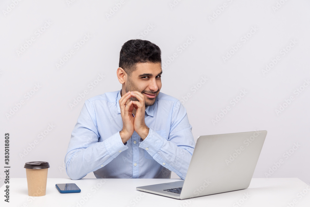 Sneaky scheming businessman sitting office workplace with laptop on desk, clasping hands and smirking mysteriously, having tricky cunning plan in mind. indoor studio shot isolated on white background Sneaky scheming businessman sitting office workplace with laptop on desk, clasping hands and smirking mysteriously, having tricky cunning plan in mind. indoor studio shot isolated on white background