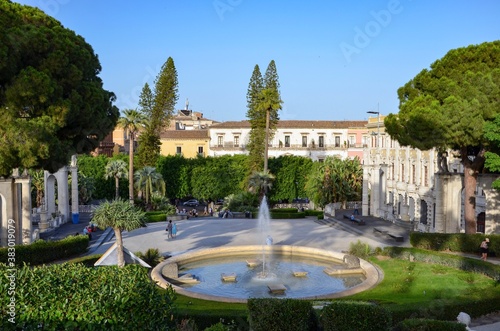 Public park called Bellini Garden (Giardino Bellini) in the city of Catania, Sicily, historic buildings on the sides, a fountain basin in the middle, mediterranean trees and plants all around, summer