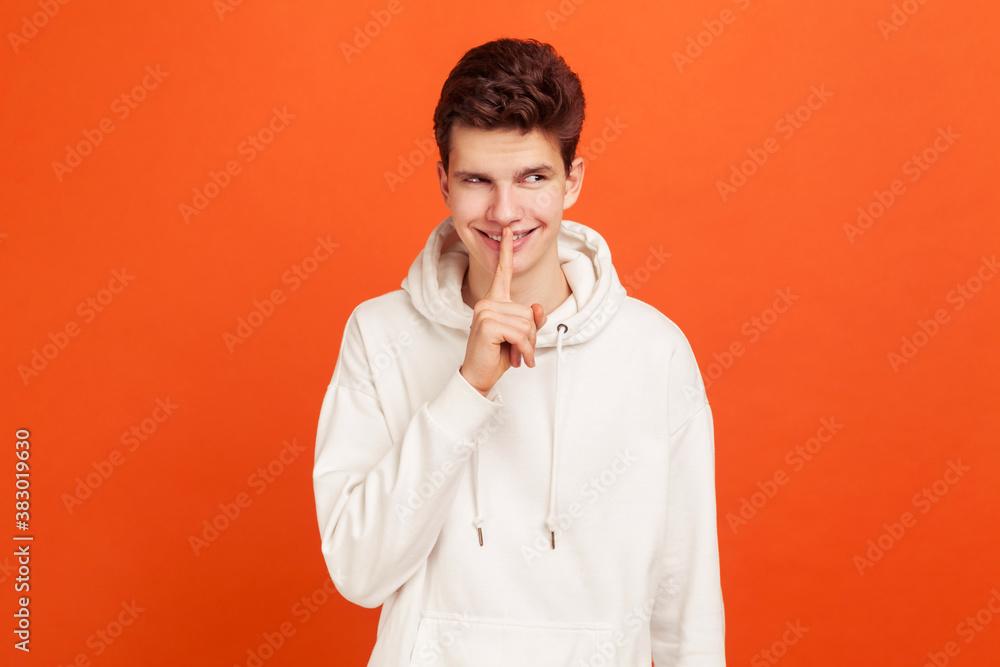 Cunning young man in casual style hoodie showing silence gesture, holding finger near lips and smiling, keep calm knowing secrets. Indoor studio shot isolated on orange background Cunning young man in casual style hoodie showing silence gesture, holding finger near lips and smiling, keep calm knowing secrets. Indoor studio shot isolated on orange background