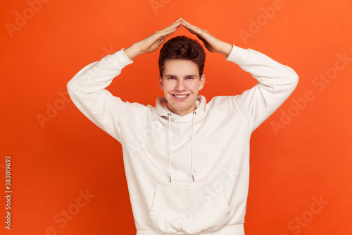 Happy smiling man in casual style sweater holding hands above head showing roof, housing on credit for youth, social security. Indoor studio shot isolated on orange background © khosrork