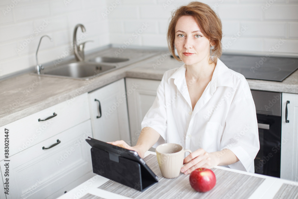 Happy beautiful woman with cup of coffee or tea using laptop in quarantine lockdown in the kitchen in the white shirt.