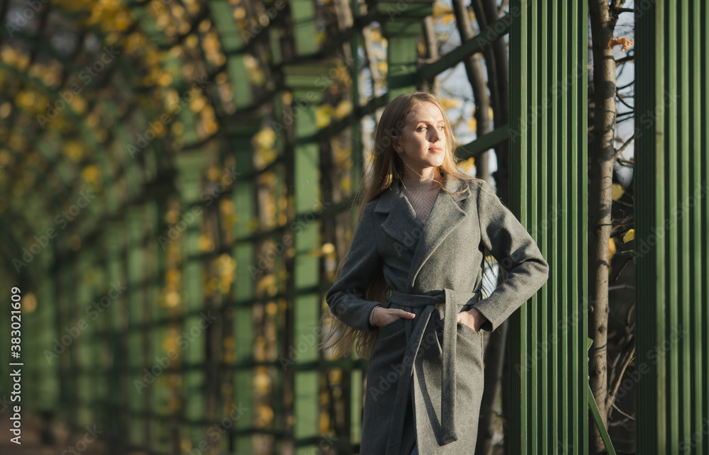 Fototapeta premium A beautiful young woman in a gray coat stands near a green fence in an autumn park. An attractive woman looks to the side pensive and a little sadly. Melancholic autumn mood. Fall season.