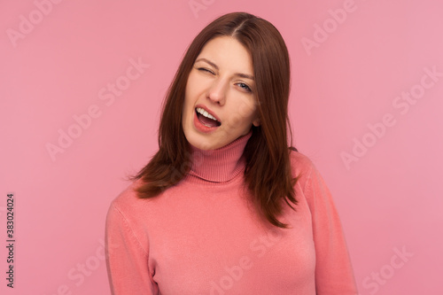 Closeup portrait of coquettish funny brunette woman in pink sweater winking looking at camera, cheering up with wink. Indoor studio shot isolated on pink background