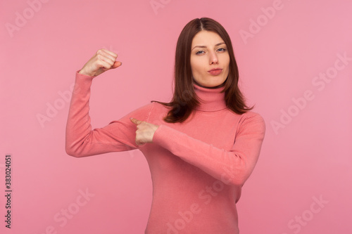 Strong self confident brunette woman in pink sweater showing her arm muscles raising hand, free and independent, emancipation. Indoor studio shot isolated on pink background