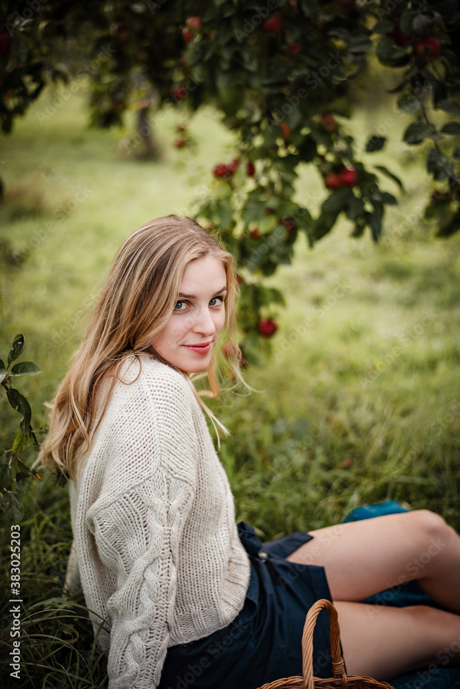 Blonde woman is sitting under a tree full of red apples. She is wearing a cozy sweater and a skirt, enjoying warm autumn evening in a garden Blonde woman is sitting under a tree full of red apples. She is wearing a cozy sweater and a skirt, enjoying warm autumn evening in a garden
