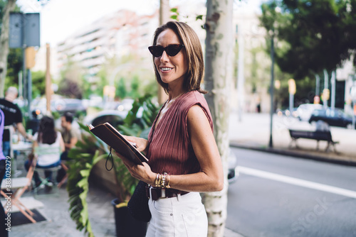 Half length portrait of beautiful trendy dressed business woman standing with planner on city street smiling, charming caucasian female in stylish sunglasses looking at camera holding notebook
