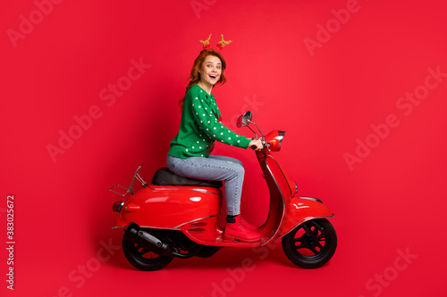 Photo portrait side view of woman riding scooter wearing green jumper deer toy headwear isolated on vivid red colored background