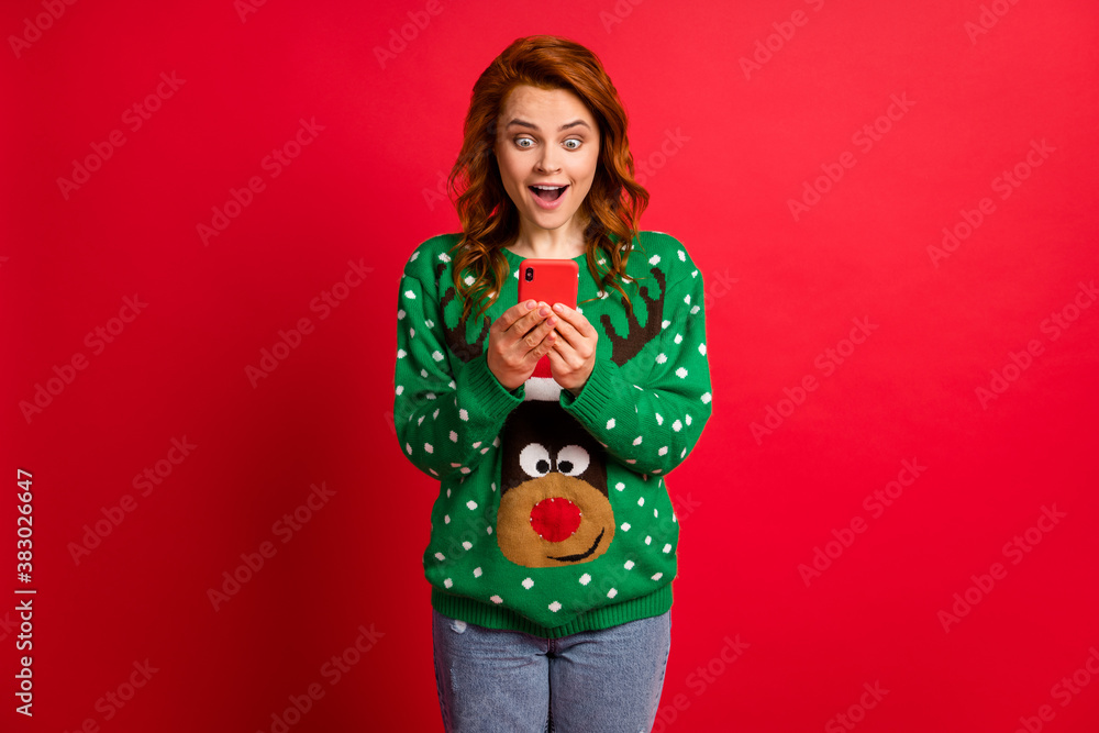 Photo portrait of excited girl with open mouth holding phone in two hands wearing ugly green sweater isolated on vivid red colored background Photo portrait of excited girl with open mouth holding phone in two hands wearing ugly green sweater isolated on vivid red colored background