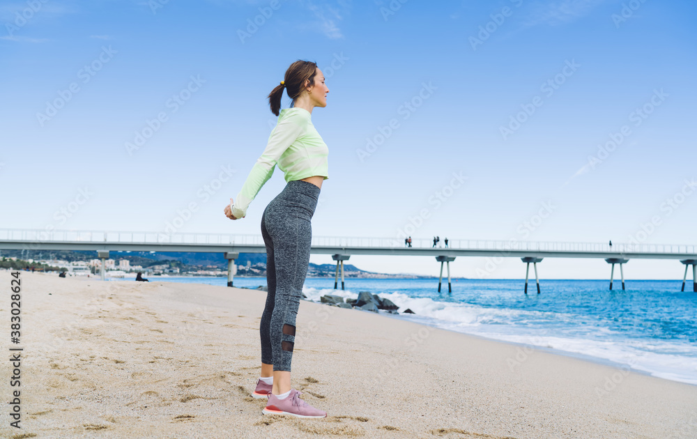 Strong female trainer in tracksuit for morning jogging stretching body muscles during warm up exercising at coastline,flexible fit girl enjoying outdoors pilates during leisure time at serene seashore