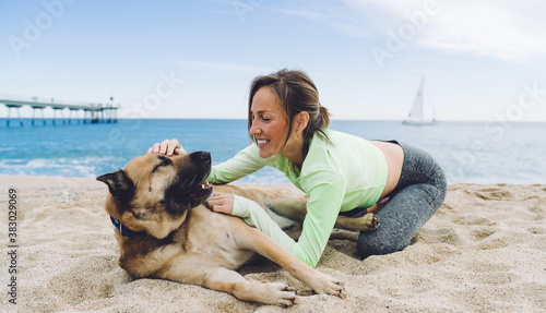 Joyful fit woman having fun with adorable mongrel dog with dark muzzle while spending leisure time at coastline seashore, playful female runner in active tracksuit stroking cute doggie pet © BullRun