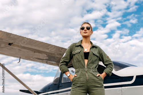 Fototapete Woman pilot wearing overall and sunglasses, standing next to a private plane on blue sky background