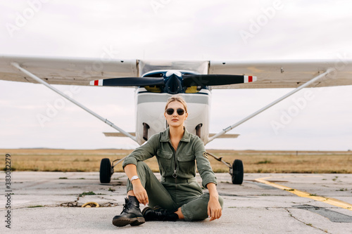 Fényképezés Confident woman pilot wearing overall and sunglasses, sitting in front of an airplane