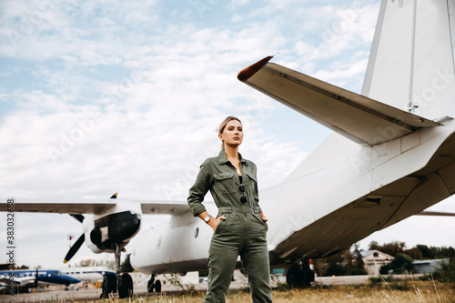 Confident woman pilot wearing overall, standing in front of an airplane with hands in pockets Fototapeta