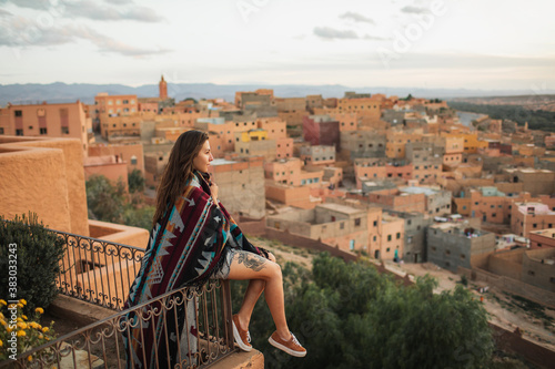 Woman sitting on roof terrace with aerial view of old arabian town Boumalne. Covered with traditional moroccan cape. Hippie style, tattoo on naked leg.