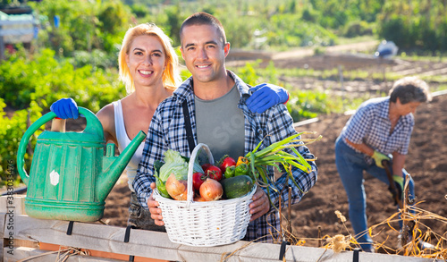 Portrait of cheerful young man and woman with basket full of freshly picked vegetables posing at garden