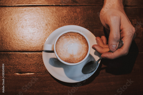 Taza de café con leche en un plato blanco sobre una mesa de madera con una mano sujetando una cuchara de plata 