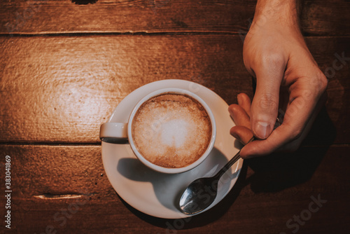 Taza de café con leche en un plato blanco sobre una mesa de madera con una mano sujetando una cuchara de plata 