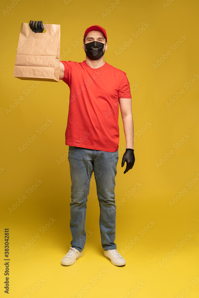 Fototapeta premium Man in red cap, t-shirt in protective mask and gloves giving fast food order isolated on yellow background. Male employee courier hold empty paper packet with food