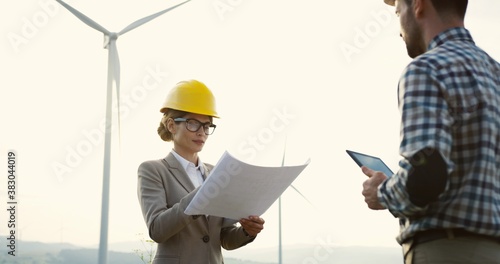 Caucasian attractive wiman boss in a helmet witj a document in hands checking the work of the worker at the windmills turbines farm.