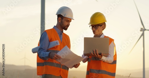 Caucasian young handsome engineer in a helmet holding some documents and drawings while having a discussion with his female boss with a laptop computer at the windmills renewable energy station.