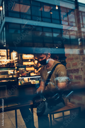 Young man sitting in coffee shop at store front in the city center in the evening, wearing the face mask to avoid virus infection and to prevent the spread of disease in time of coronavirus