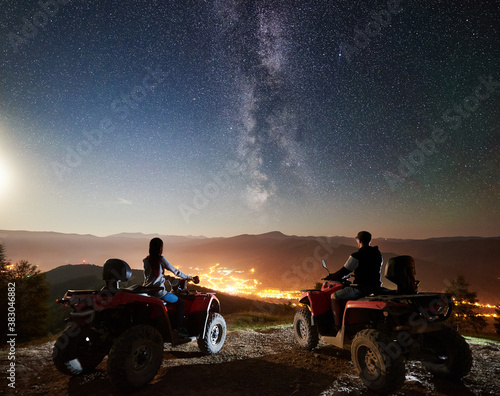 Back view of man and girl trevelers riding on atv quad motorbikes on the top of mountain, romantic couple enjoying beautiful view of night sky full of stars, Milky way, luminous city on background photo