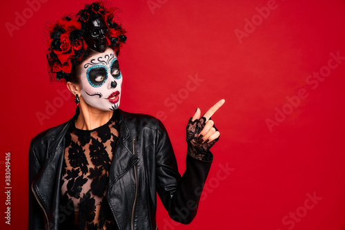 Young woman with sugar skull makeup with a wreath of flowers on her head and skull and black gloves points to a free space. isolated on red background. concept of Halloween or Calavera Catrina