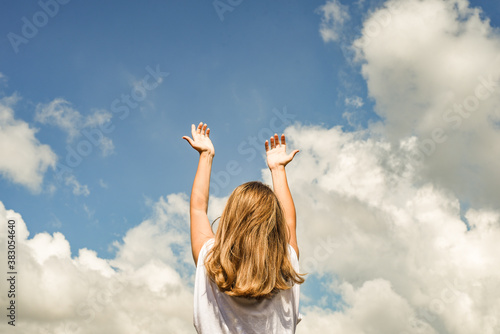 a woman or girl stands with her back to the camera and stretches her hands to the sky. The girl raised her hands up against the blue sky
