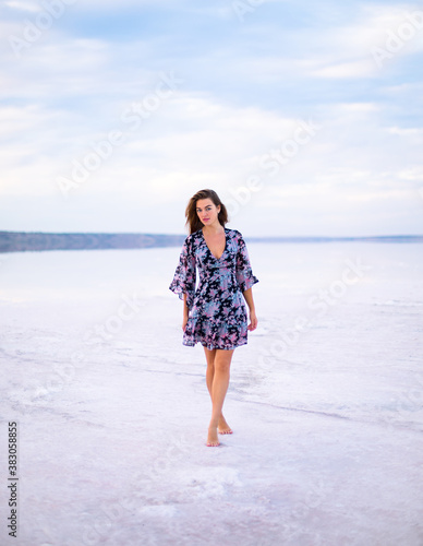 beautiful girl with brown hair in a black dress with a color pattern walks on a salty pink still water lake at a vanilla sunset